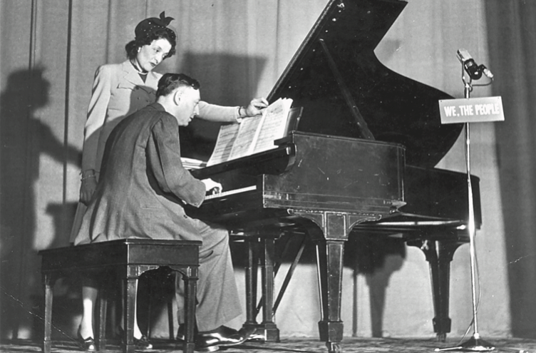 An image of a music therapist turning the page of sheet music for a patient playing the piano