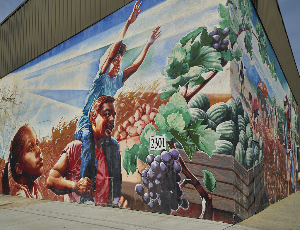 A mural of two children, one on their father's back, with crates of produce and a grape vine