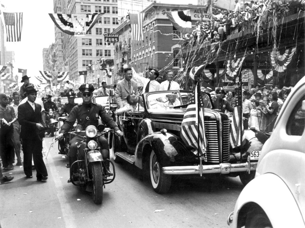 A black and white photo of Howard Hughes riding in a motorcade in a parade celebrating his new record in aviation 