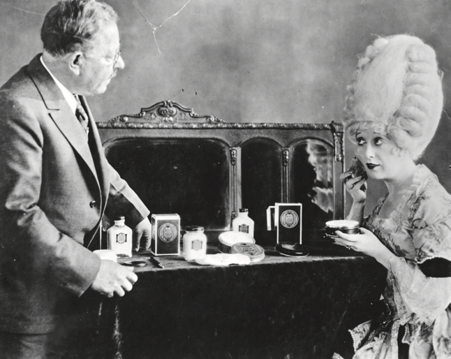 Max Factor and actress Mabel Normand at a vanity while Mabel applies makeup.