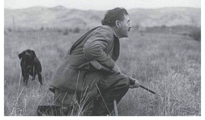 A side profile of Ernest Hemingway crouching with a gun as Bullet the dog runs up behind him